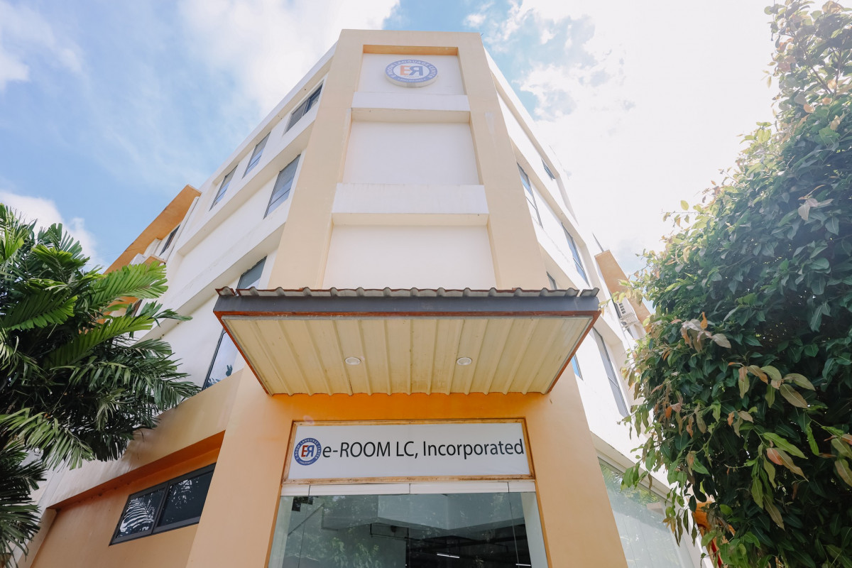 巴科羅 EROOM 語言學校的米黃色校舍大樓,入口雨棚上掛著 e-ROOM LC, Incorporated 招牌,旁邊有綠樹