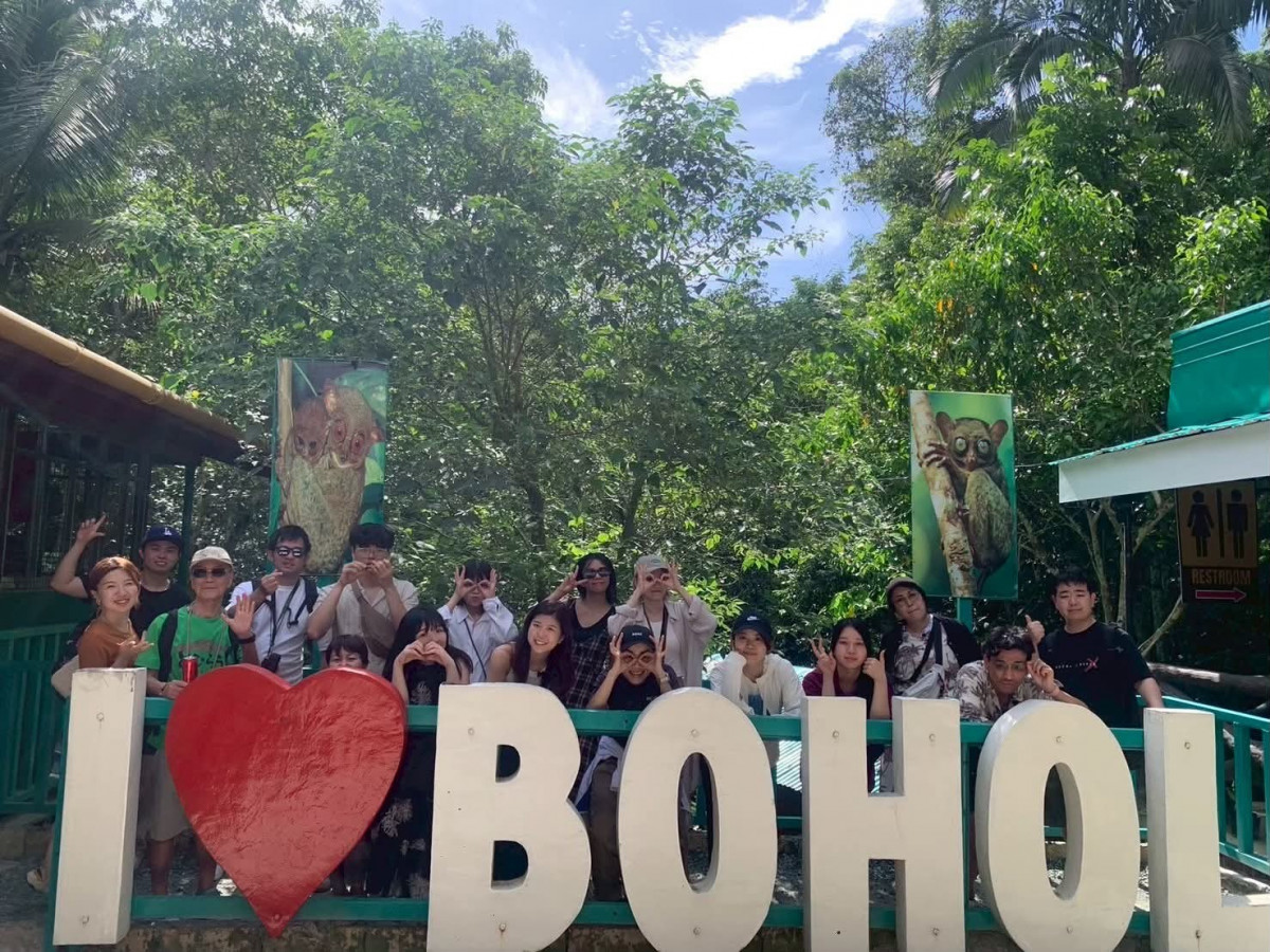 一群學生擠在薄荷島 I ♥ BOHOL 白色大字牌後面合照，背後是綠樹和眼鏡猴看板
