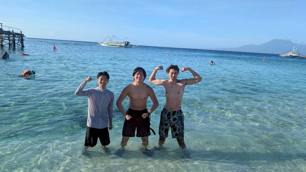 三位男學生站在清澈見底的淺海裡擺出秀肌肉的姿勢，遠方海面停著白色螃蟹船