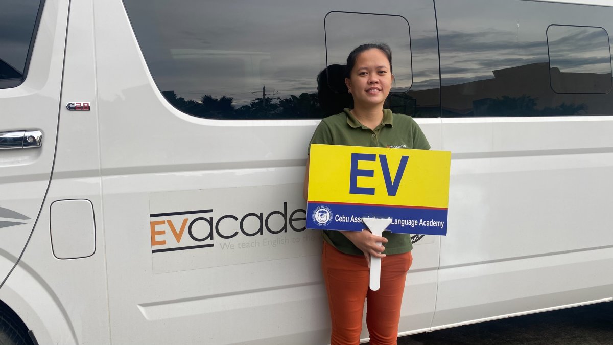 宿霧 EV 語言學校的接機人員舉著黃色 EV 接機牌,站在車身印著 EVacademy 的白色廂型車旁