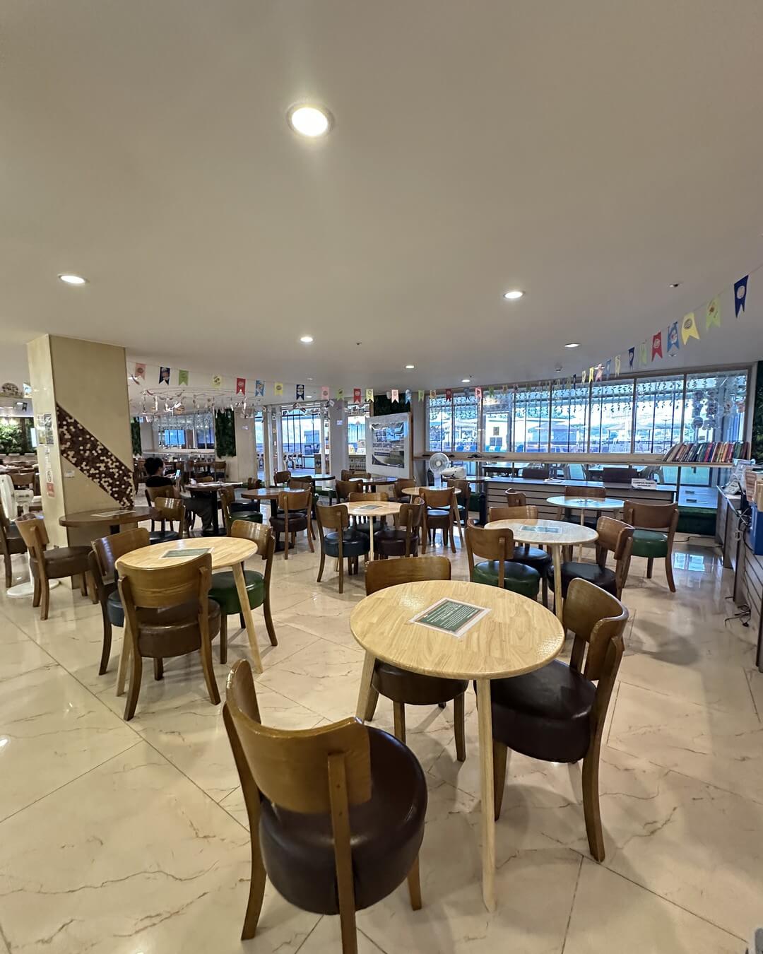 宿霧麥克坦島 SMEAG ENCANTO 校區的學生餐廳,一張張木頭圓桌配皮面椅,天花板下掛著彩色三角旗,窗邊有一排書架