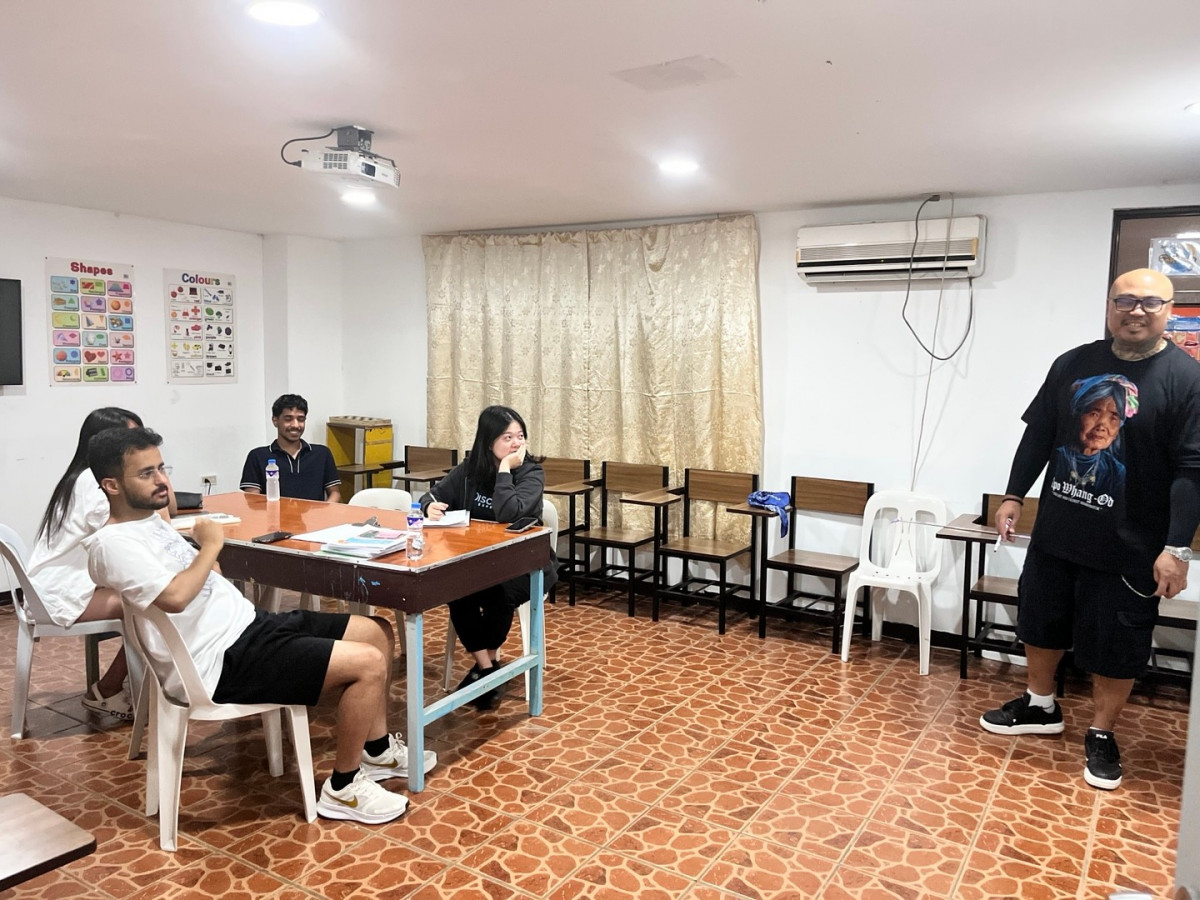 克拉克 GS 語言學校的外師團體課，老師拿著白板筆站在教室一側，幾位成人學生圍著長桌坐，牆上貼著英文海報