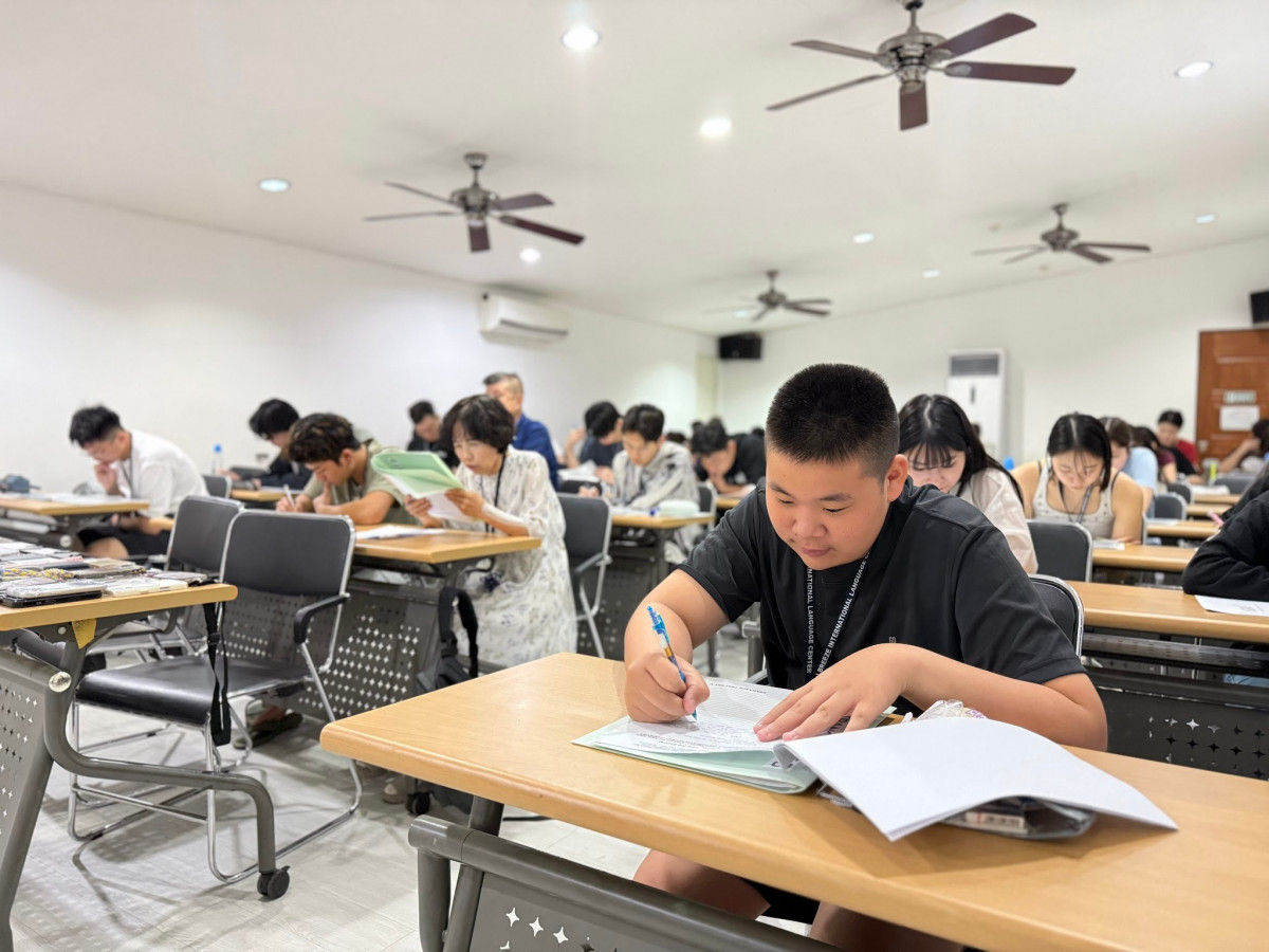 語言學校的大教室裡，新生坐在一排排長桌前低頭寫入學測驗卷，天花板掛著好幾支吊扇