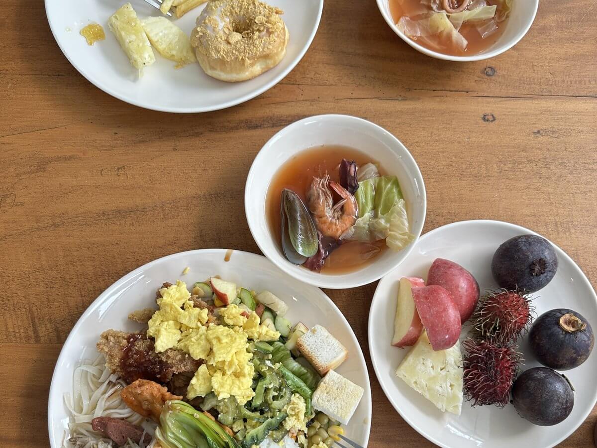 宿霧 EV 語言學校午餐自助吧夾好的一份午餐,盤裡有麵條、炒蛋、青菜、麵包丁和炸物,旁邊一碗有蝦和淡菜的湯,另一盤是蘋果、紅毛丹和山竹