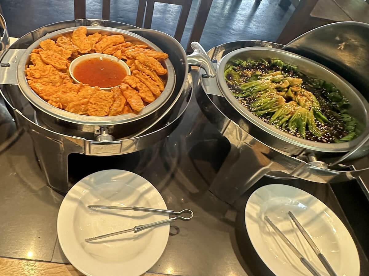 宿霧 EV 語言學校餐廳的自助吧檯,保溫餐爐裡一鍋金黃炸物配沾醬、一鍋淋醬撒芝麻的青江菜,前面擺著白瓷盤和夾子
