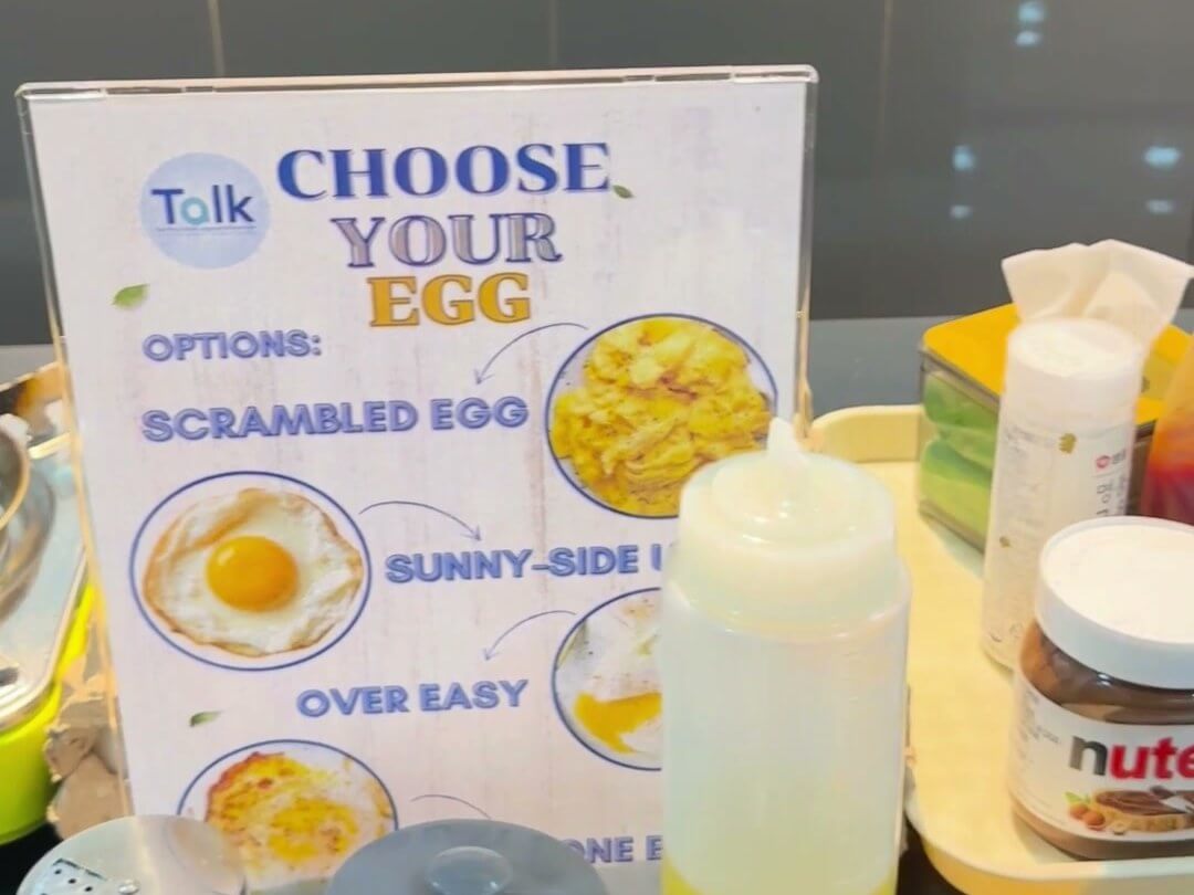 克拉克 TALK 語言學校新校區餐廳的早餐檯,立牌寫著 CHOOSE YOUR EGG,列出炒蛋、太陽蛋等做法,前面有調味罐和醬料瓶
